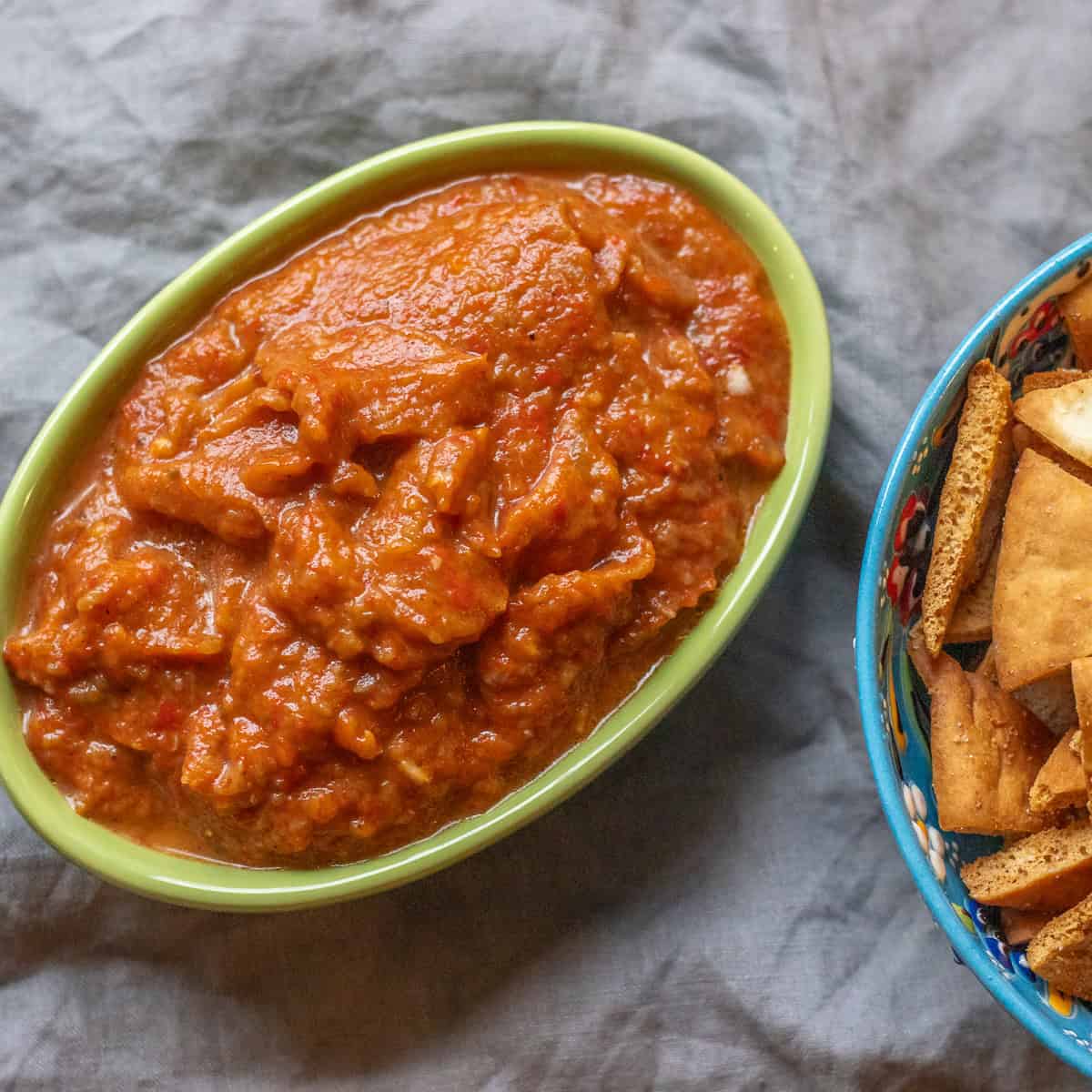 Ajvar in a green oval dish beside a patterned bowl of pita chips.